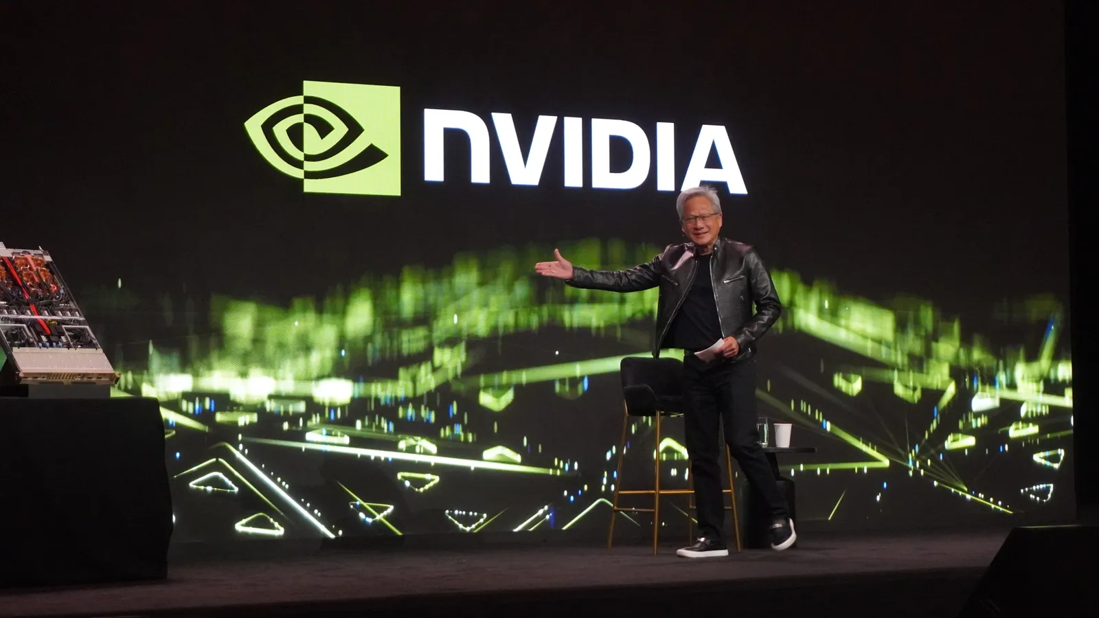 Photo of a keynote stage. A speaker in a black leather jacket gestures towards a giant screen carrying the Nvidia logo over green abstract graphics, with a server unit on a plinth to one side.
