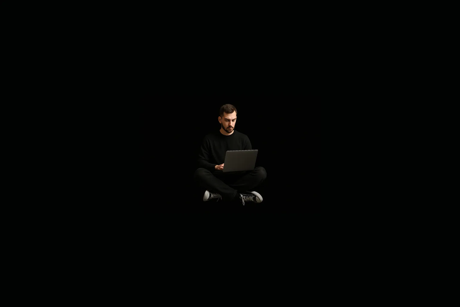 A person sitting cross-legged with a laptop, lit alone in an otherwise entirely black frame.