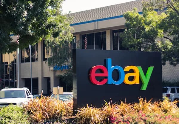 Photo of the eBay headquarters sign, the multicoloured ebay wordmark mounted on a black plinth in a landscaped bed, with the low office building and a US flag behind it.