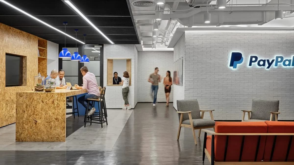 Photo of a modern PayPal office interior. An illuminated PayPal logo sits on a white brick wall on the right, above armchairs and an orange bench sofa. On the left, people work at a plywood counter under blue pendant lamps, and others walk through the corridor between.