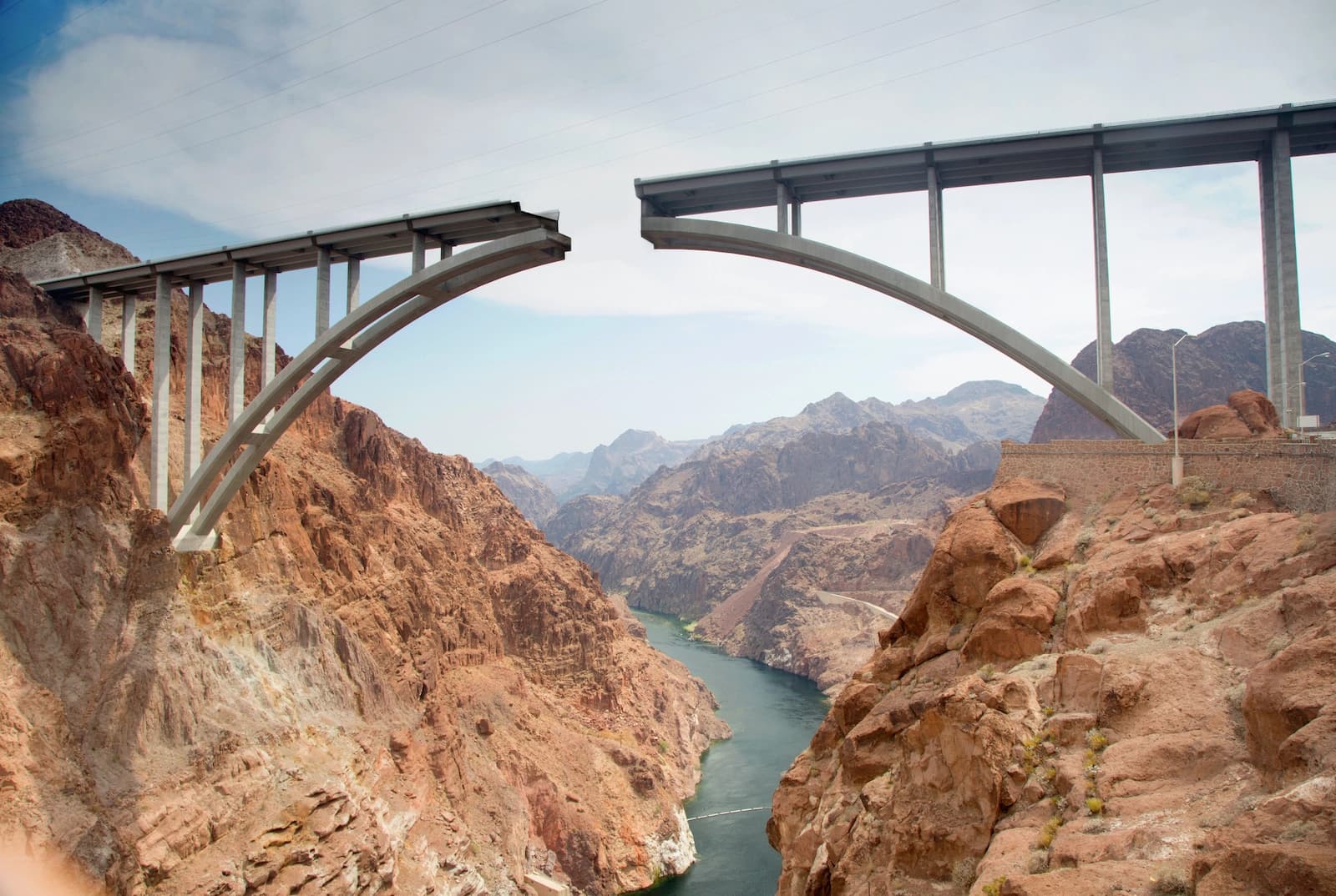 An unfinished bridge across a desert canyon, its two arched halves reaching toward each other with a gap still open above the river below.