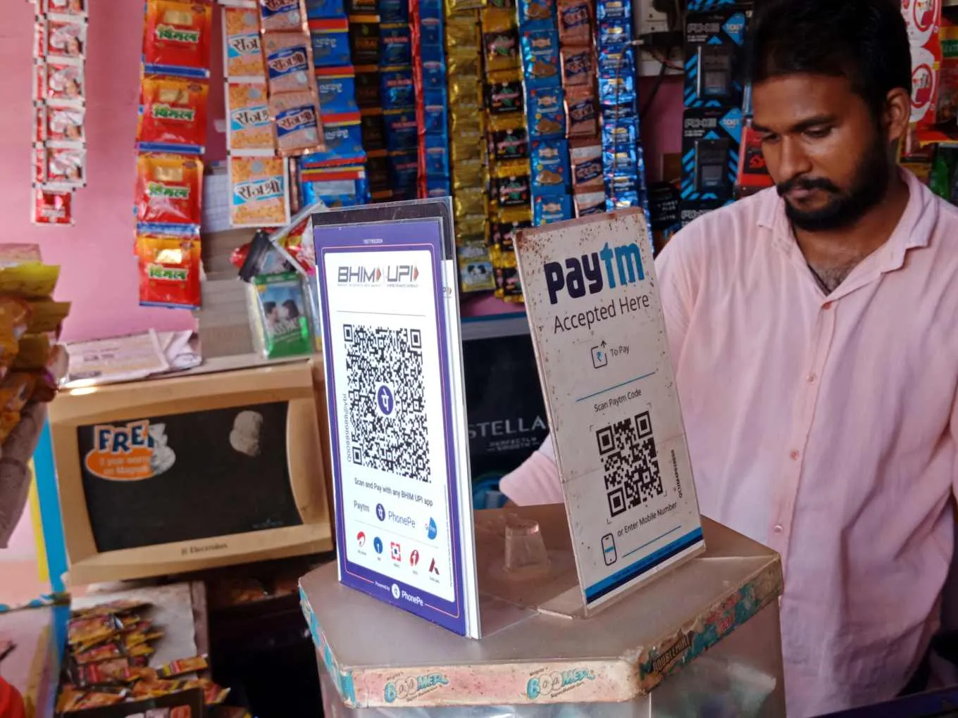 Photo of a small Indian shop counter with two QR code stands side by side. One is a BHIM UPI card reading "Scan and Pay with any BHIM UPI app" listing Paytm, PhonePe and other apps and powered by PhonePe; the other reads "Paytm accepted here, scan Paytm code, or enter mobile number". The shopkeeper stands behind them, with strips of snack sachets hanging above the counter.