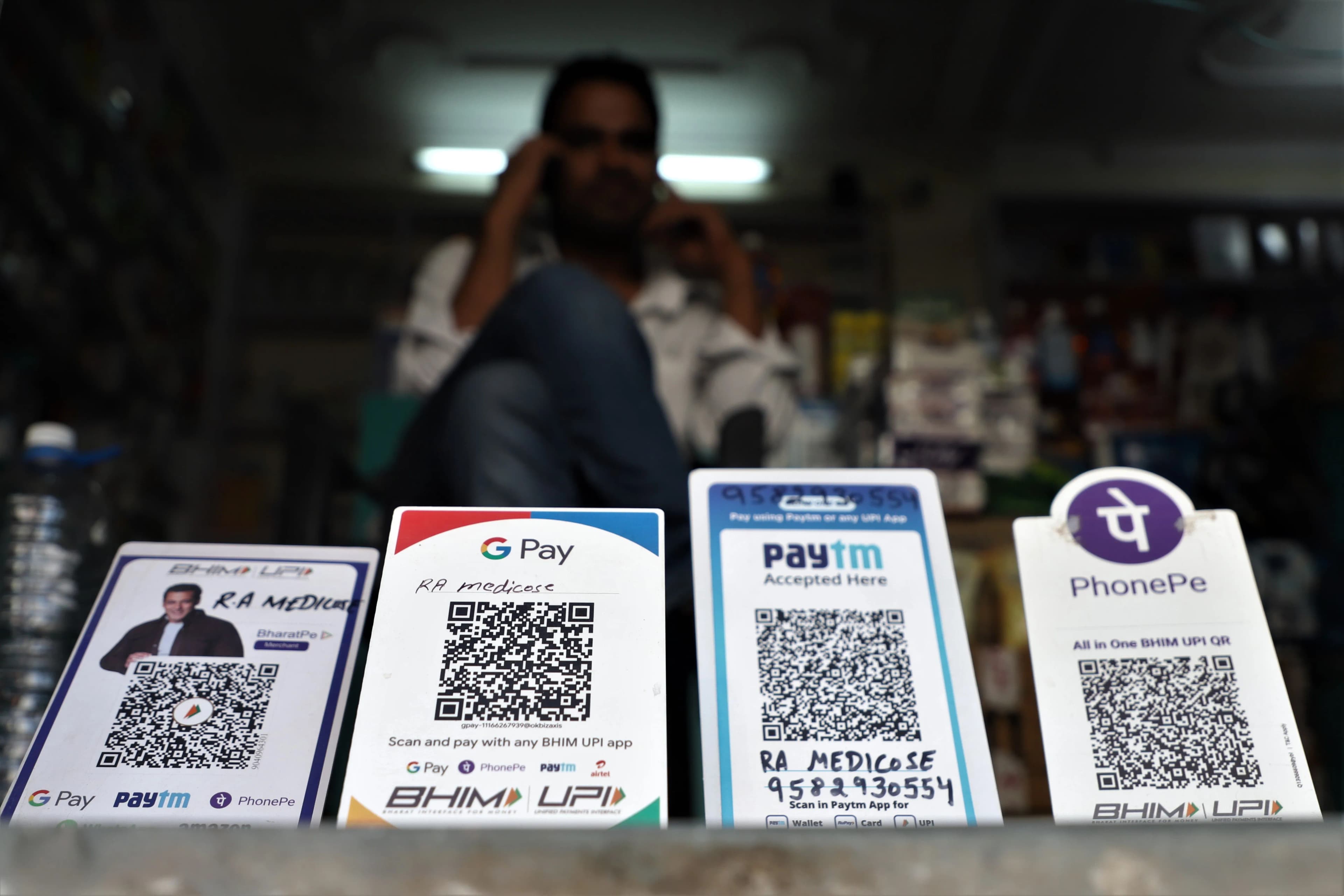 Photo of four payment QR code stands lined up on a shop counter, for BHIM UPI via BharatPe, Google Pay, Paytm and PhonePe, each carrying the merchant name RA Medicose and a phone number, with the PhonePe card labelled "All in One BHIM UPI QR". The shopkeeper is out of focus behind them, on the phone.
